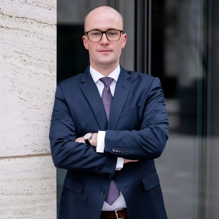 Rechtsanwalt Raphael Griemert in Anzug mit Brille, siegesgewiss vor modernem Gebäude.