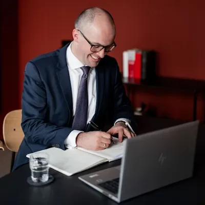 Rechtsanwalt Raphael Griemert im Anzug mit lächelndem Gesicht vor neutralem Hintergrund schreibt etwas.